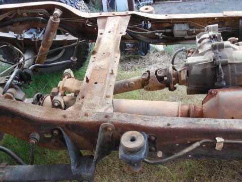 1993 Ford Bronco - Rusty boxed truck frame with the body off, viewed over the transmission crossmember: a two-piece driveshaft with U-joints runs back from th…