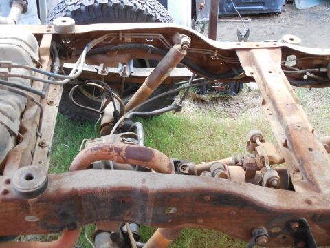 1993 Ford Bronco - Rusty bare truck frame on grass with the body removed, showing new black body-mount bushings on the rails, a steering damper and drag link…