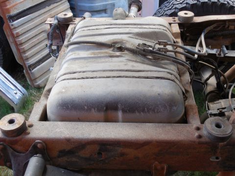 1993 Ford Bronco - Looking down into a rusty truck frame with the body off, a dusty steel fuel tank sits between the rails held by two straps, with fuel lines…