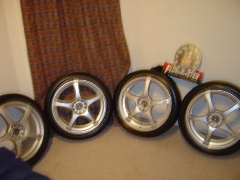 1998 Ford Taurus - A collection of four aftermarket wheels and tires are displayed indoors, accompanied by a Flowmaster sticker and a gauge.