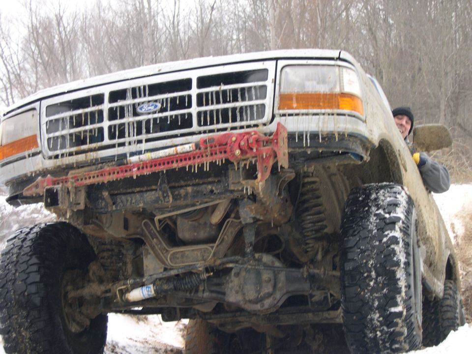 Mud-covered white Ford 4x4 nose-high on snowy trail with red Hi-Lift jack on front bumper