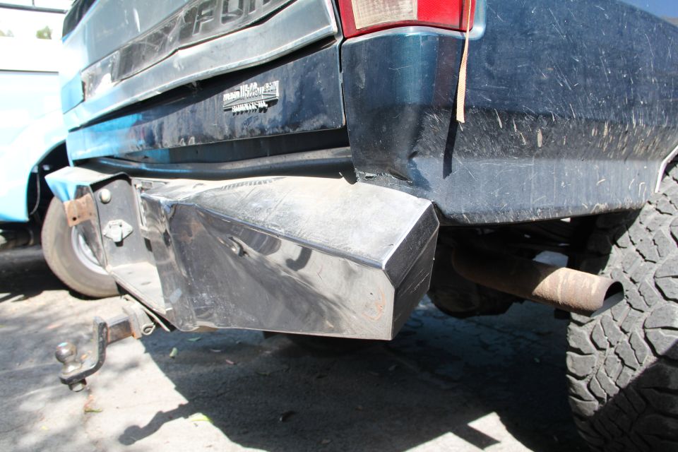 Dark blue Ford F-250 truck rear with custom metal bumper and tow hitch. Visible tire and exhaust pipe