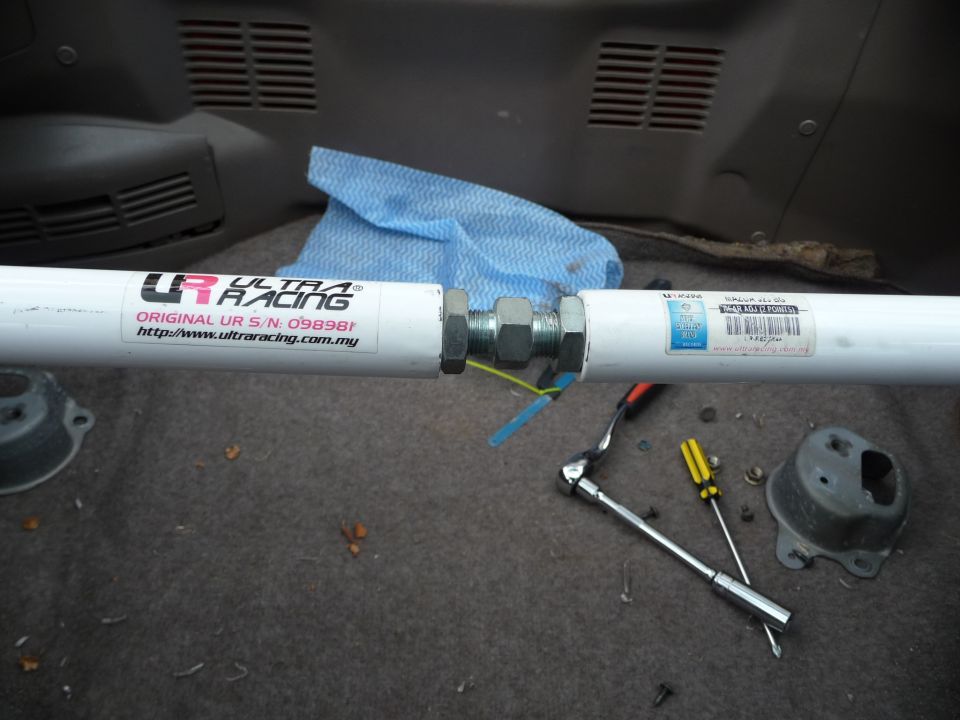 White Ultra Racing strut bar with serial number and website visible, lying on carpet with tools