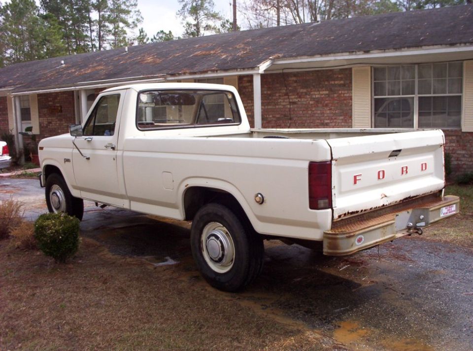 White Ford regular-cab long-bed pickup, rear three-quarter view, parked on gravel by a brick house