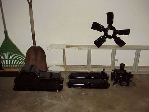 1989 Ford Bronco - Various black engine parts including an oil pan, valve covers, water pump, and fan blade are laid out on the floor.