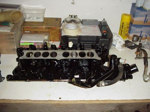 1989 Ford Bronco - An engine intake manifold sits on a surface, surrounded by boxes, tape, and other automotive parts.