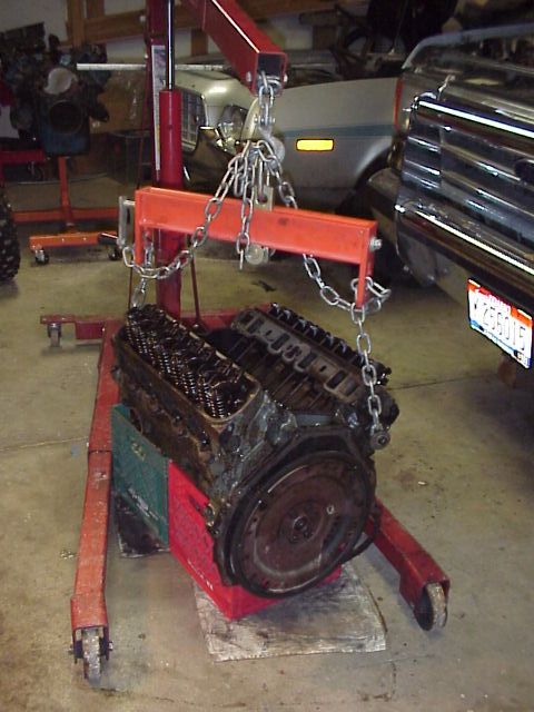 1989 Ford Bronco - An engine is suspended by a red engine hoist, resting on crates, with other vehicles nearby.