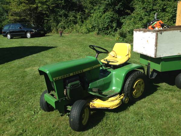 Green John Deere lawn tractor with yellow seat, towing a green trailer with a white box containing a chainsaw