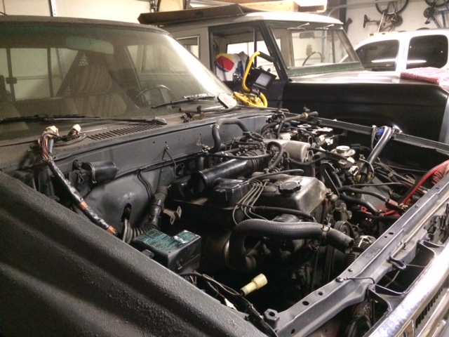 Engine bay of a truck with the hood up, showing the engine and wiring. Another vehicle is visible behind it