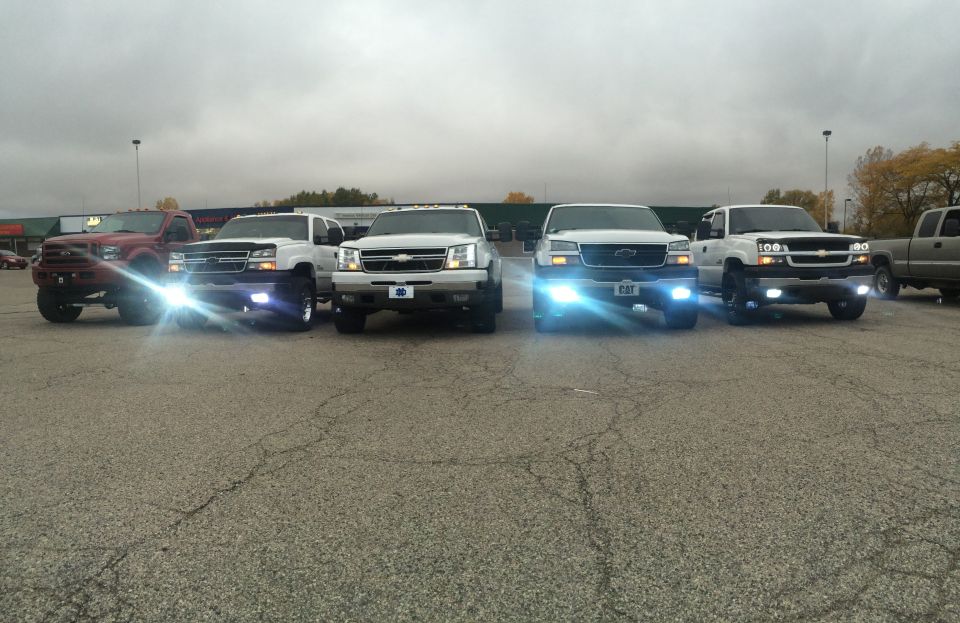 Four Chevrolet Silverado trucks parked in a lot, three white and one red, with bright headlights