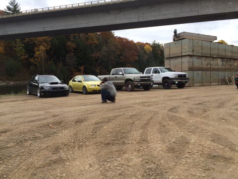 2006 Chevrolet Silverado 2500 - A group of trucks and cars are parked outdoors under a large bridge, with a photographer crouching to take a picture.