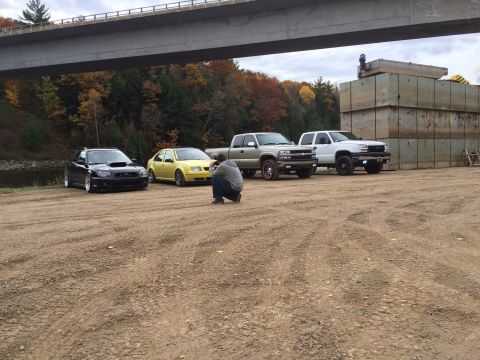 2006 Chevrolet Silverado 2500 - A photographer kneels to take a picture of several vehicles parked on a dirt lot near a bridge.