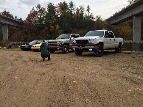 2006 Chevrolet Silverado 2500 - A white 2006 Chevrolet Silverado 2500 pickup truck is parked on a dirt lot with other vehicles and bridges in the background.