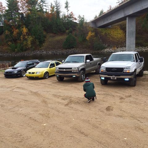 2006 Chevrolet Silverado 2500 - A group of trucks and cars are parked near a river with a bridge overhead. A person is taking a photo of the vehicles.