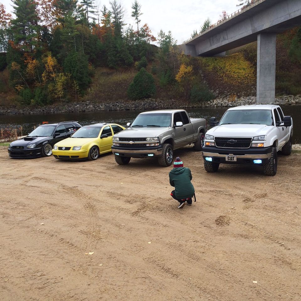 Four vehicles parked by a river under a bridge, person crouching to take photo