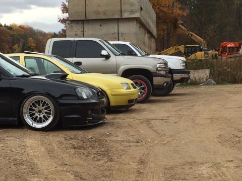 2006 Chevrolet Silverado 2500 - A lineup of modified cars and a pickup truck are parked outdoors with construction equipment in the background.