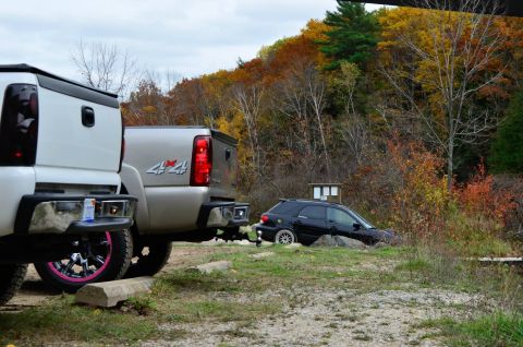 2006 Chevrolet Silverado 2500 - Two pickup trucks are parked on gravel, with a black SUV partially off-road in the background amidst autumn foliage.