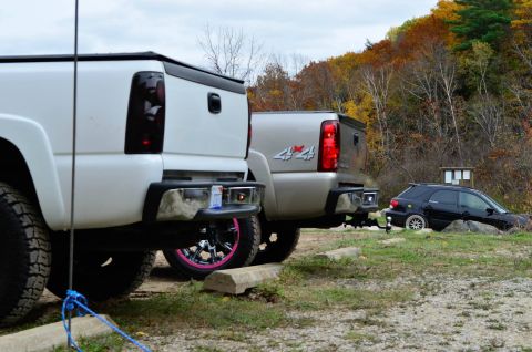 2006 Chevrolet Silverado 2500 - Rear view of a white Chevrolet Silverado 2500 and a silver pickup truck, with a black SUV in the background, parked on a dirt lot with fall…