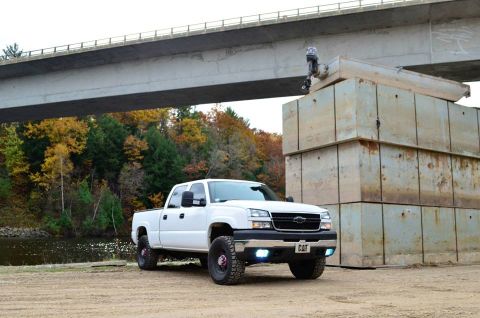 2006 Chevrolet Silverado 2500 - A white Chevrolet Silverado pickup truck is parked on a dirt lot near a body of water, with a bridge overhead and stacked concrete blocks t…