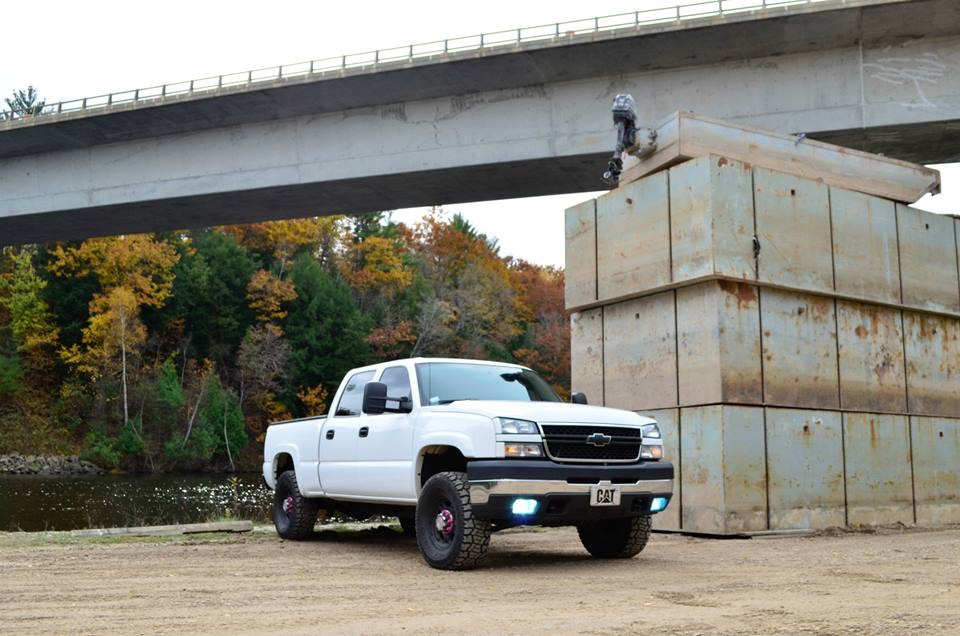 White Chevrolet Silverado pickup truck with aftermarket wheels and CAT badge on the grille, parked on dirt near a body of water and under a bridge