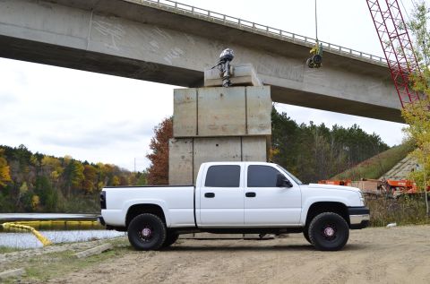 2006 Chevrolet Silverado 2500 - A white pickup truck is parked on a dirt lot next to a body of water, with a bridge overhead and construction equipment in the background.