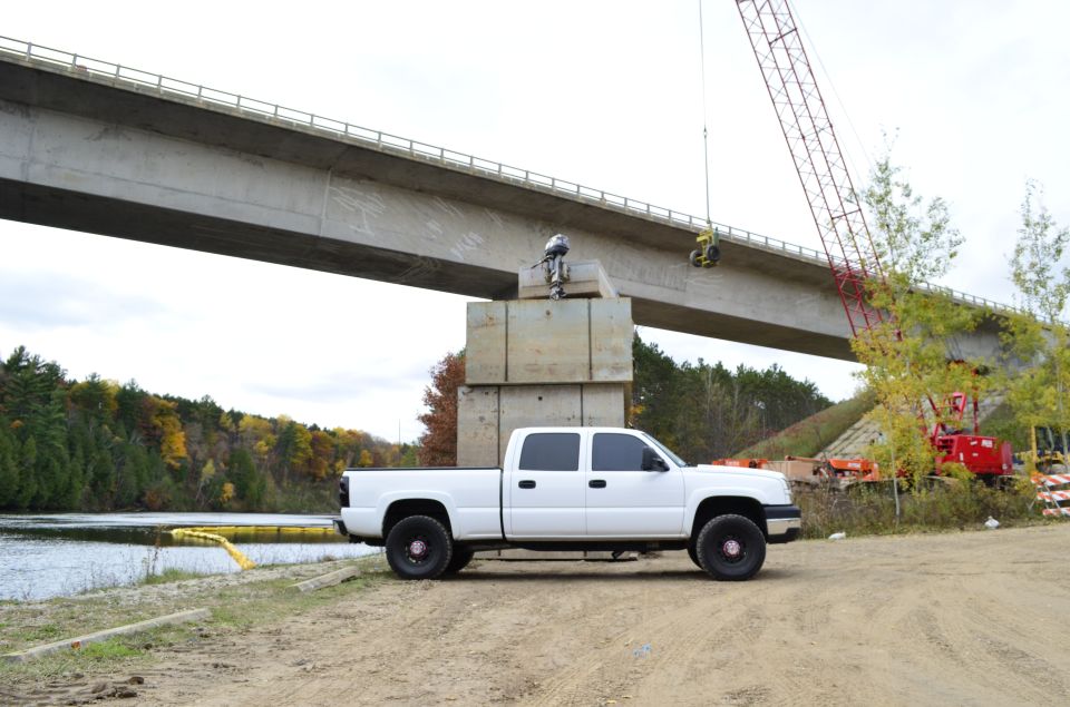 White 2006 Chevrolet Silverado 2500 parked on dirt near a river, under a bridge under construction with a crane