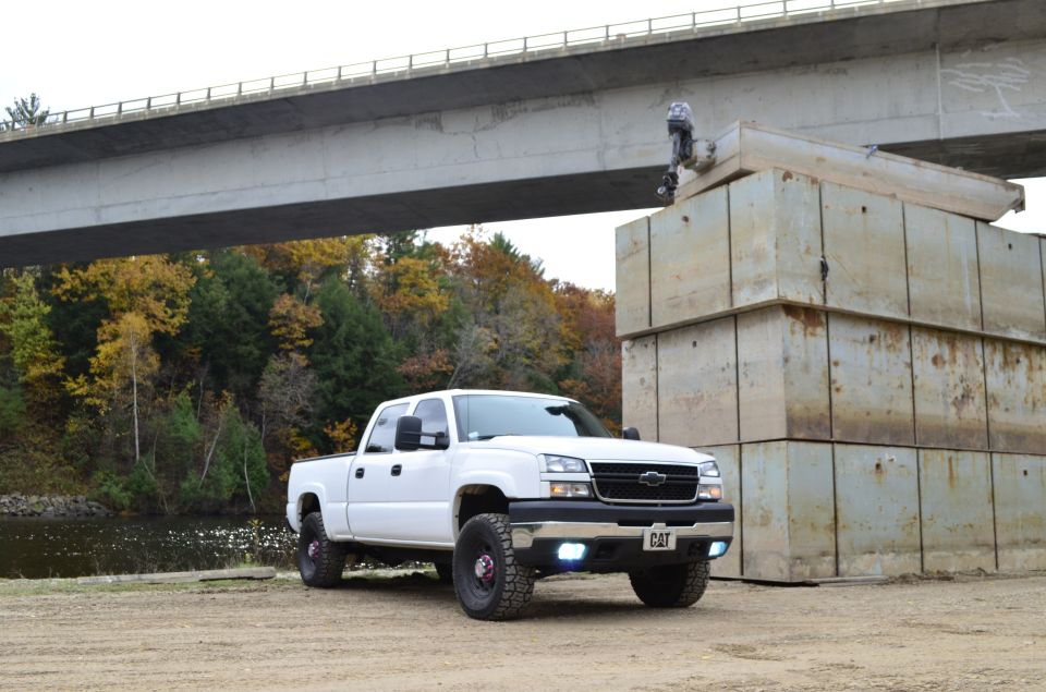 White pickup truck with aftermarket wheels and CAT badging on the grille, parked on dirt near a bridge and concrete blocks