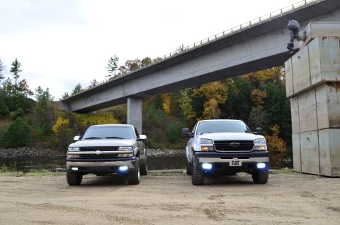 2006 Chevrolet Silverado 2500 - Two Chevrolet Silverado trucks are parked on a dirt lot under a large concrete bridge with autumn trees in the background.