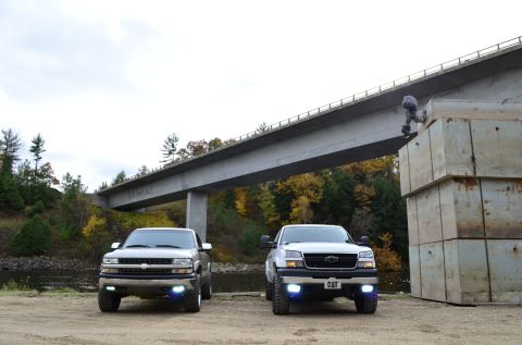 2006 Chevrolet Silverado 2500 - Two Chevrolet Silverado 2500 trucks are parked side-by-side under a large concrete bridge with autumn foliage in the background.