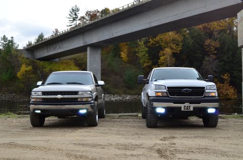 2006 Chevrolet Silverado 2500 - Two Chevrolet Silverado trucks are parked side-by-side under a bridge with autumn foliage in the background.
