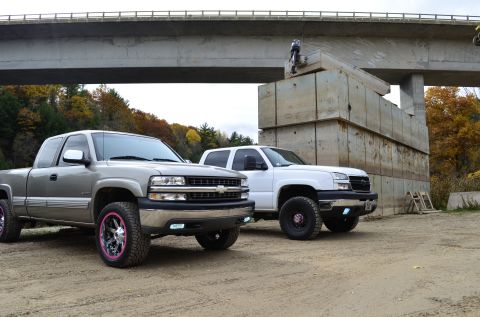2006 Chevrolet Silverado 2500 - Two pickup trucks are parked on a dirt lot near a concrete structure and a bridge overhead.