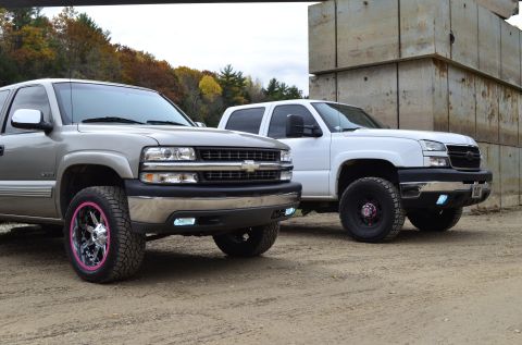 2006 Chevrolet Silverado 2500 - Two Chevrolet Silverado trucks are parked outdoors, with one in silver and the other in white, against a backdrop of concrete blocks and tr…