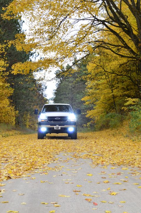 2006 Chevrolet Silverado 2500 - A pickup truck drives down a road lined with trees in peak fall foliage, with the ground covered in fallen leaves.