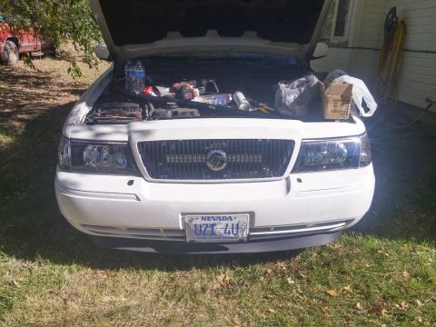 2000 Mercury Grand Marquis - The front of a white 2000 Mercury Grand Marquis is shown with its hood open, revealing a cluttered engine bay.