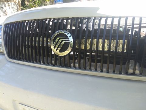 2000 Mercury Grand Marquis - Close-up of the front grille on a white 2000 Mercury Grand Marquis, the Mercury emblem and auxiliary lighting.