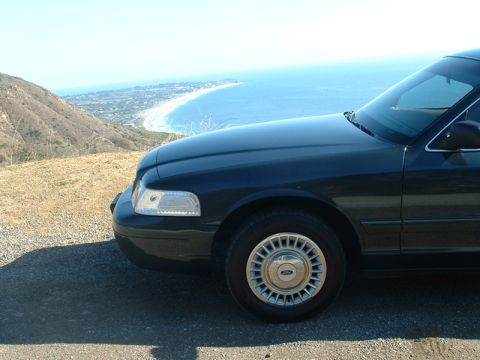 1998 Ford Crown Victoria - Front fender and headlight of a dark green Ford sedan parked at a hilltop overlook above the ocean.