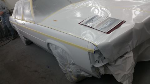 1991 Ford LTD Crown Victoria - A white sedan is covered in plastic and tape, likely in a body shop, with a binder resting on its hood.