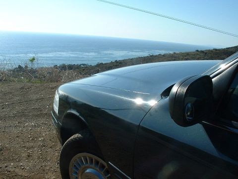 1998 Ford Crown Victoria - Front fender, hood, and side mirror of a dark green sedan parked at a hilltop overlook by the ocean.