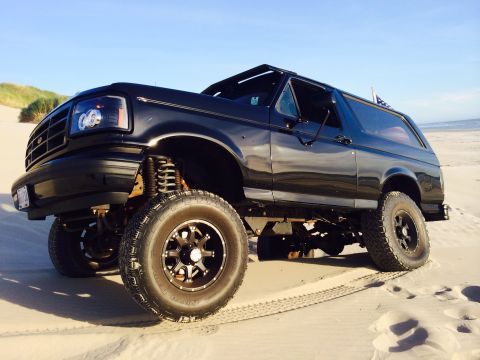 1992 Ford Bronco - A lifted black 1992 Ford Bronco is parked on a sandy beach under a clear blue sky.