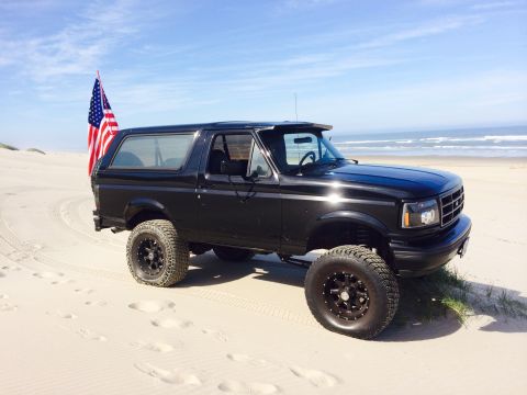 1992 Ford Bronco - Black 1992 Ford Bronco parked on a sandy beach with the ocean in the background.