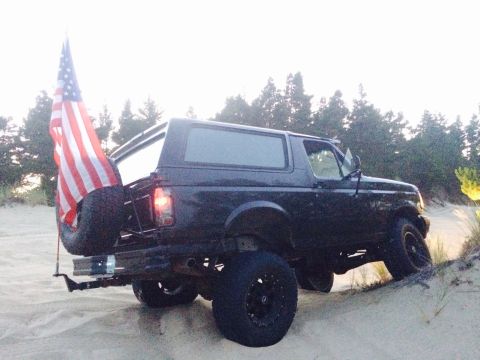 1992 Ford Bronco - This 1992 Ford Bronco is shown from the rear, driving on a sandy terrain with an American flag displayed.