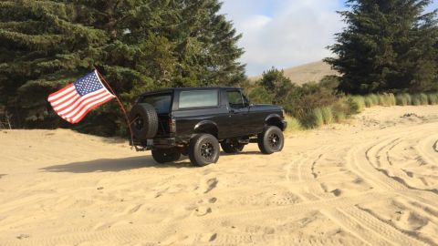 1992 Ford Bronco - This black 1992 Ford Bronco is parked on a sandy dune, flying an American flag from its rear spare tire mount.
