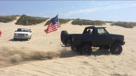 1992 Ford Bronco - Two Ford Broncos drive on sand dunes under a clear sky, one with an American flag.