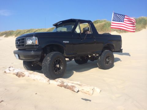 1992 Ford Bronco - A black 1992 Ford Bronco is parked on a sandy dune with an American flag flying in the background.