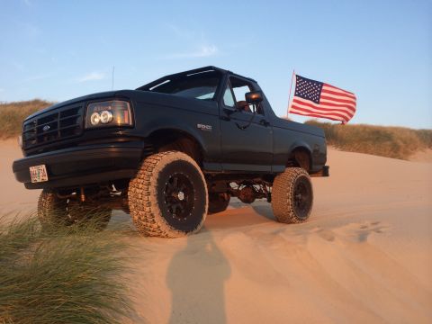 1992 Ford Bronco - Black 1992 Ford Bronco with an American flag flies on sand dunes under a clear sky.