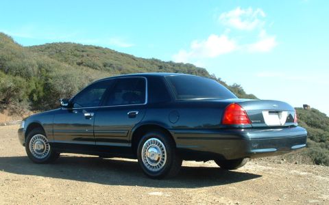 1998 Ford Crown Victoria - dark green Ford Crown Victoria sedan sits on a dirt clearing among brushy hills, shown from the rear three-quarter angle.