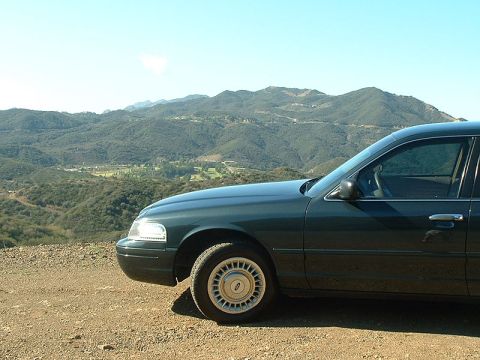 1998 Ford Crown Victoria - front quarter of a dark green sedan parked on a dirt overlook above a green valley with mountains in the distance.