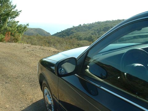 1998 Ford Crown Victoria - Close view of the side mirror and front fender of a dark sedan parked on a dirt road overlooking wooded hills.