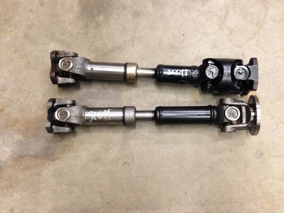 Two drive shafts, one silver and one black, laid out on a concrete floor. Text is written on both