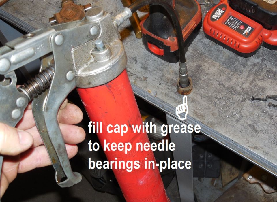 Hand holding red grease gun on workbench with tool text overlay about greasing needle bearings